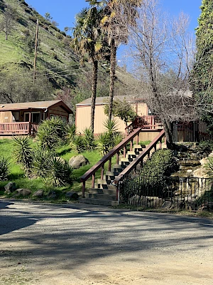 The image shows a small, rustic building with a staircase leading up, surrounded by trees and shrubs on a sunny day, against a hillside.